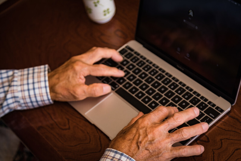 ノート型パソコンのキーボードに年配男性の手が添えられている画像
